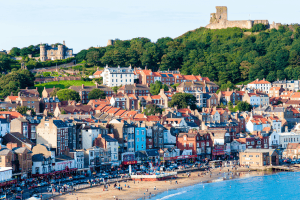 Scarborough south bay a beach with coastal buildings all different colours, blue, yellow, white. Scarborough Castle can be seen in the background a stone tower on top of a hill over the town.