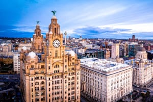 Aerial view of Liverpool cityscape featuring historic buildings including the Royal Liver Building with its prominent clock tower under a partly cloudy sky.